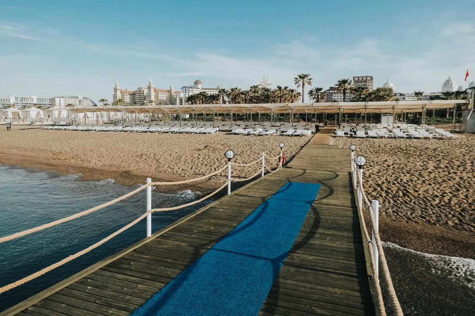 Houten pier met touwreling over het water bij Saturn Palace Resort, met het strand en de ligbedden erachter
