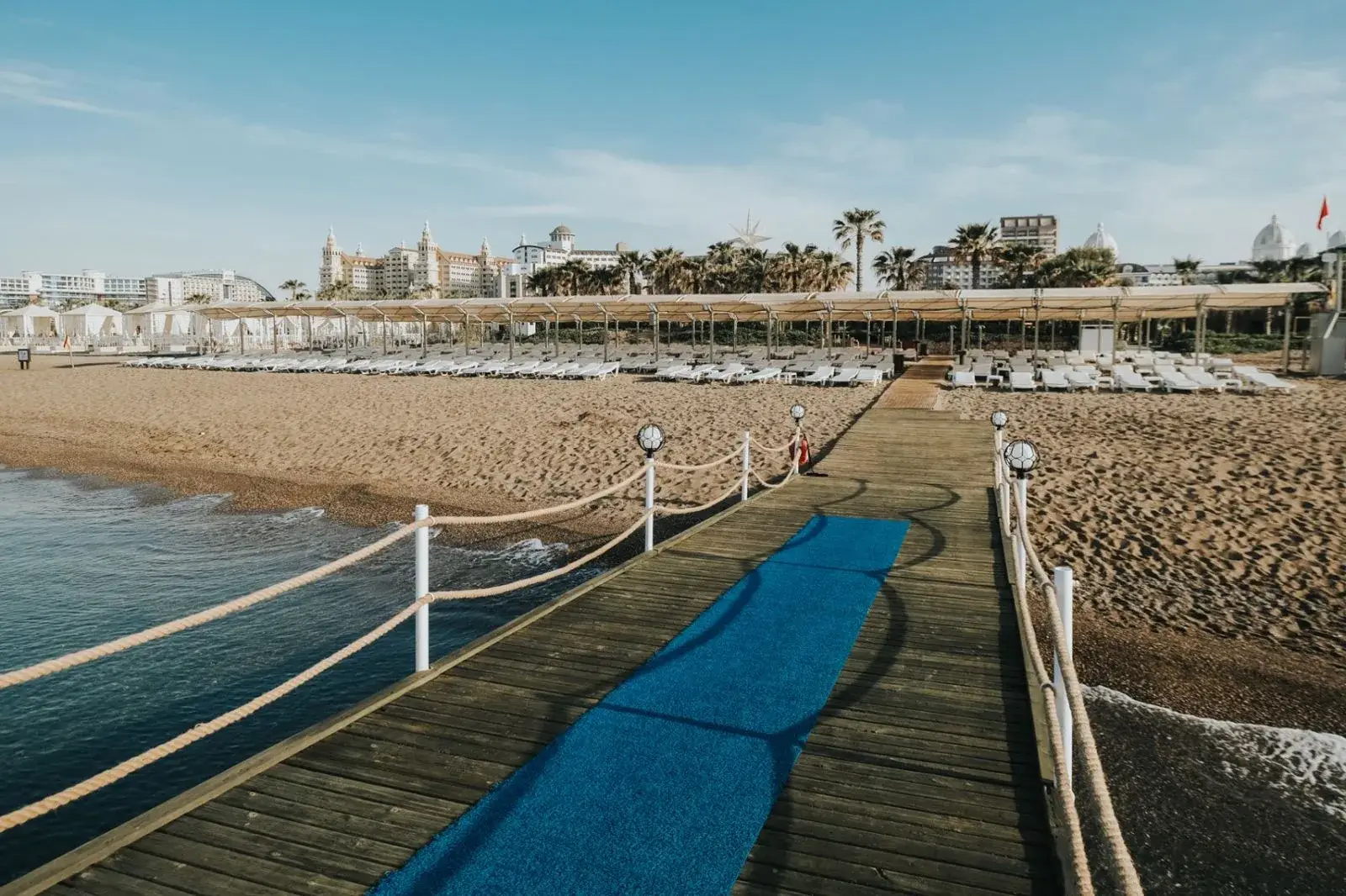 Houten pier met touwreling over het water bij Saturn Palace Resort, met het strand en de ligbedden erachter